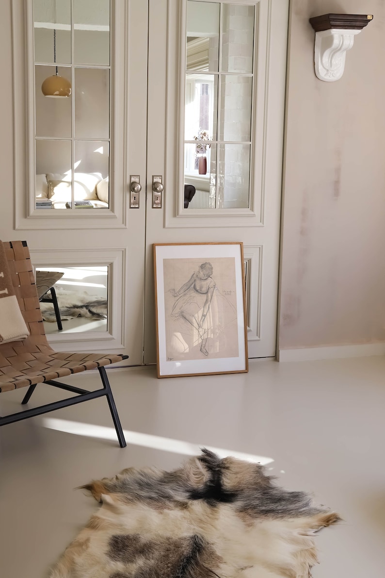 May include: A framed pencil sketch of a seated figure, leaning against a wall. The artwork is displayed on a light-colored floor, with a natural fur rug in the foreground. A woven leather chair and a set of double doors with glass panels are also visible.