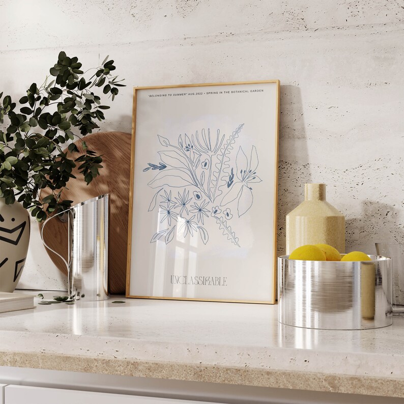 Op de afbeelding: Een ingelijst drukwerk met een gouden lijst en een witte achtergrond. De print toont een blauwe lijntekening van bloemen en bladeren. De tekst "BELONGING TO SUMMER, AUG 2019 - SPRING IN THE BOTANICAL GARDEN" en "UNCLASSIFIABLE" zijn op de print gedrukt.