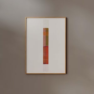 Op de afbeelding: Een ingelijst kunstwerk met een kleurrijk geweven wandkleed met een lange, dunne vorm. Het wandkleed is gemaakt van verschillende gekleurde vierkanten en rechthoeken, met een overheersend oranje en rode kleurenschema. Het wandkleed hangt aan een lichtgrijze muur.