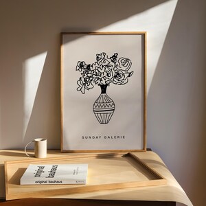 May include: A black and white line drawing of a bouquet of flowers in a vase. The drawing is framed in a light wood frame and is hanging on a wall. The text "SUNDAY GALERIE" is printed below the drawing.