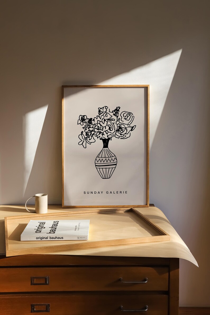 May include: A black and white line drawing of a bouquet of flowers in a vase. The drawing is framed in a light wood frame and is hanging on a wall. The text "SUNDAY GALERIE" is printed below the drawing.