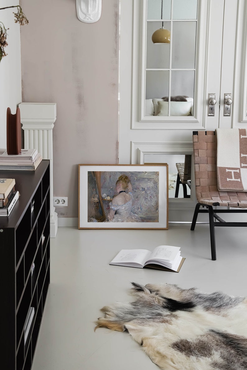 May include: A framed painting of a woman in a white dress sitting on a chair. The painting is hanging on a light pink wall in a room with a white rug and a wooden chair.
