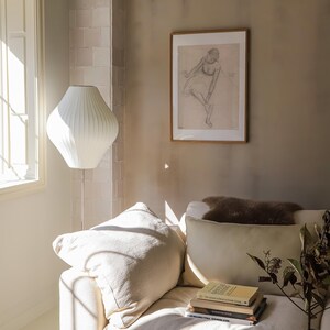 May include: A cozy living room scene featuring a white sofa with cream-colored pillows and a stack of books. A white floor lamp with a unique shade stands near a window. A framed sketch hangs on the wall, adding an artistic touch.