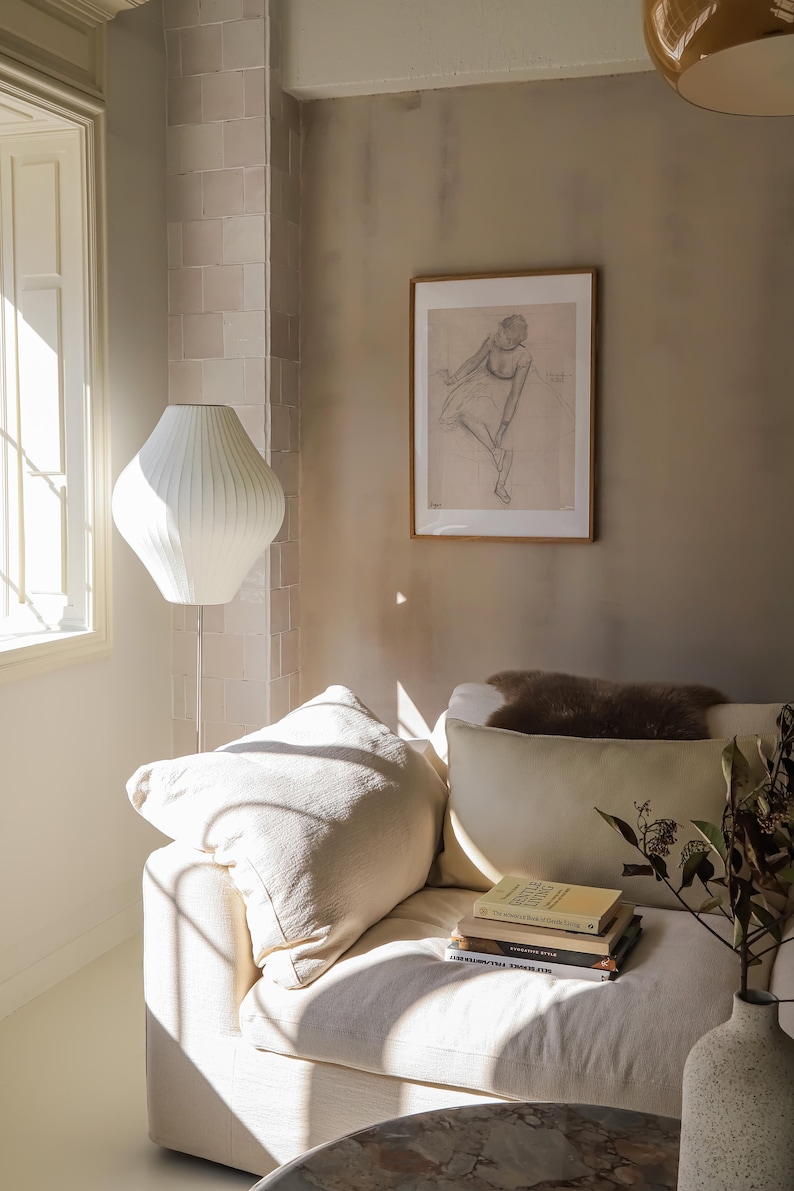 May include: A cozy living room scene featuring a white sofa with cream-colored pillows and a stack of books. A white floor lamp with a unique shade stands near a window. A framed sketch hangs on the wall, adding an artistic touch.