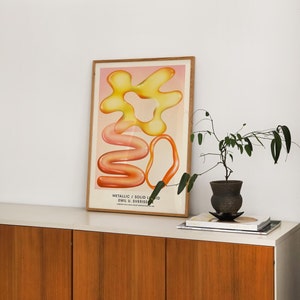 Puede incluir: Una impresi&oacute;n art&iacute;stica enmarcada con formas abstractas en tonos amarillos, naranjas y rosas. La obra, titulada "Metallic / Solid Liquid" de Emil U. Sverrisson, se exhibe sobre un mueble blanco con puertas de madera. Una peque&ntilde;a planta en maceta y libros est&aacute;n sobre el mueble.