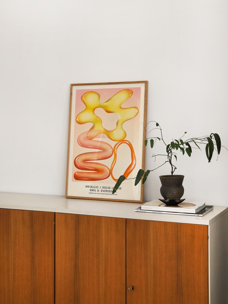 Puede incluir: Una impresi&oacute;n art&iacute;stica enmarcada con formas abstractas en tonos amarillos, naranjas y rosas. La obra, titulada "Metallic / Solid Liquid" de Emil U. Sverrisson, se exhibe sobre un mueble blanco con puertas de madera. Una peque&ntilde;a planta en maceta y libros est&aacute;n sobre el mueble.