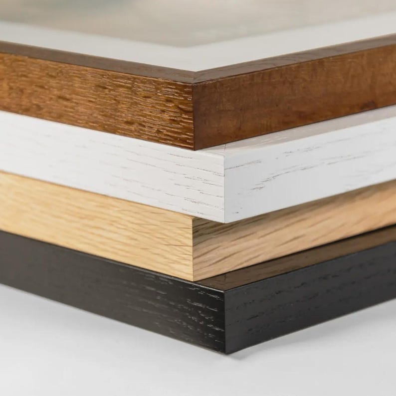 May include: A stack of four picture frames with different wood finishes. The top frame is a dark brown, followed by white, light oak, and black. The frames are stacked, showing the corner details and textures of the wood.
