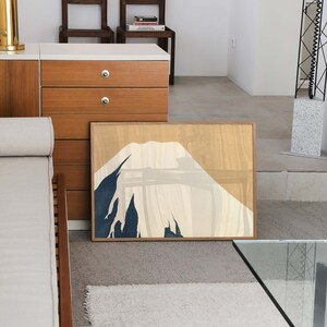 以下が含まれることがあります： 青、白、ベージュの色合いでスタイリッシュな山岳風景の額装されたプリント。山は、テクスチャのある背景でミニマルなスタイルで描かれています。