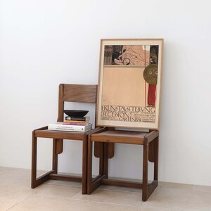 May include: Two wooden chairs with a stack of books on the left chair. A framed poster with the text "KUNSTAUSSTELLUNG SECESSION" is behind the chairs.