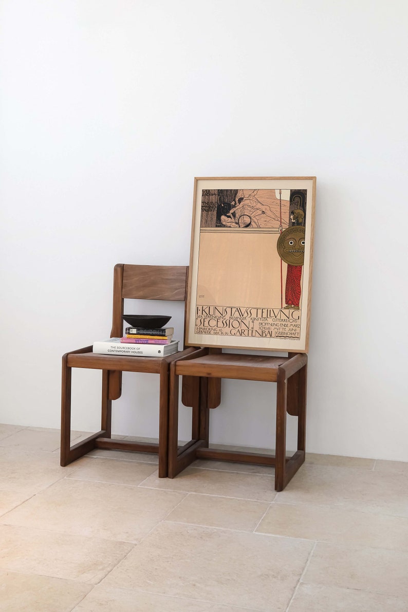 May include: Two wooden chairs with a stack of books on the left chair. A framed poster with the text "KUNSTAUSSTELLUNG SECESSION" is behind the chairs.