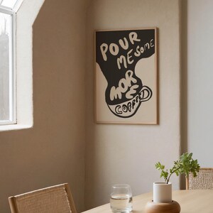 May include: A black and white poster with the text "POUR ME SOME MORE COFFEE" in a stylized font. The poster is framed in a light wood frame and is hanging on a beige wall.