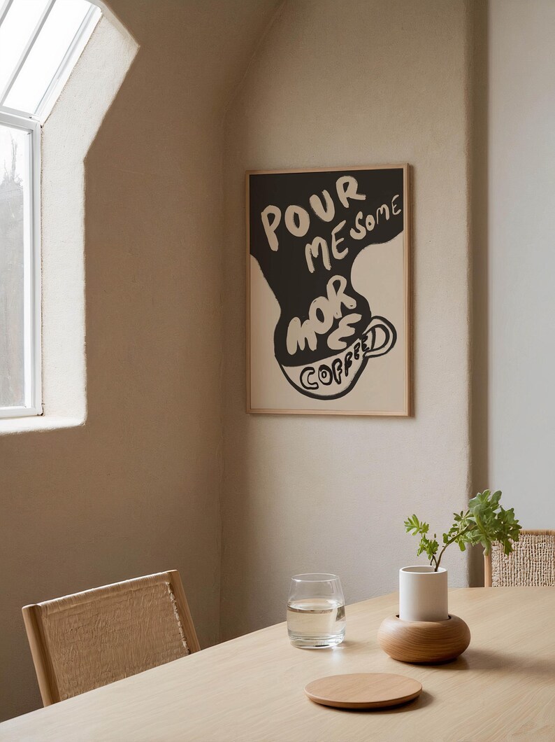 May include: A black and white poster with the text "POUR ME SOME MORE COFFEE" in a stylized font. The poster is framed in a light wood frame and is hanging on a beige wall.