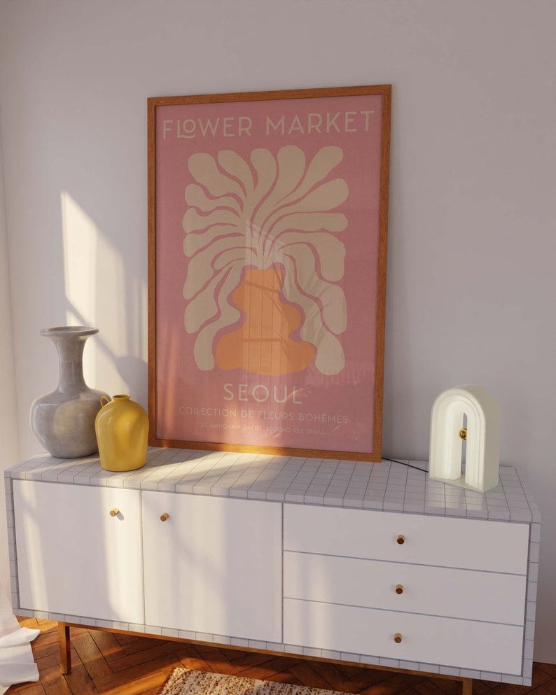 May include: A white dresser with three drawers and gold knobs. A framed poster with the text "FLOWER MARKET SEOUL COLLECTION DE FLEURS BOH&Egrave;MES" is on the dresser. A white arch-shaped lamp is on the right side of the dresser.