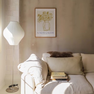 May include: A white couch with a brown throw pillow and a stack of books on the armrest. The couch is in a room with a white lamp and a framed print of a vase of flowers with the text "SUNDAY GALERIE" below the image.