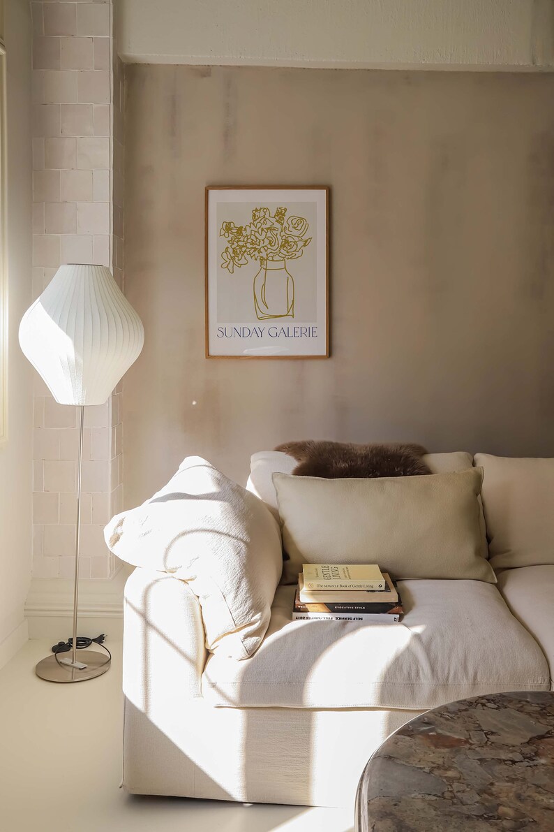 May include: A white couch with a brown throw pillow and a stack of books on the armrest. The couch is in a room with a white lamp and a framed print of a vase of flowers with the text "SUNDAY GALERIE" below the image.