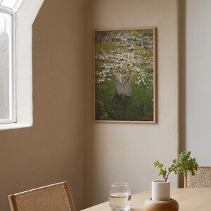 Op de afbeelding: Een ingelijst schilderij van drie vrouwen in een veld met witte bloemen. Het schilderij hangt aan een lichtbeige muur in een kamer met een houten tafel en een glas water.