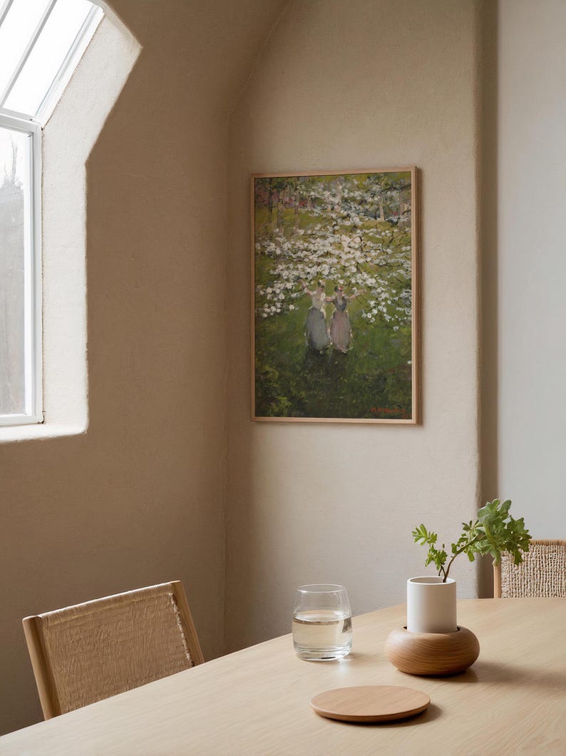 Op de afbeelding: Een ingelijst schilderij van drie vrouwen in een veld met witte bloemen. Het schilderij hangt aan een lichtbeige muur in een kamer met een houten tafel en een glas water.