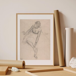 May include: Framed pencil sketch of a ballerina adjusting her shoe, with a light beige background. The artwork is in a gold frame. Rolls of paper and a wooden frame are on a light wood surface.