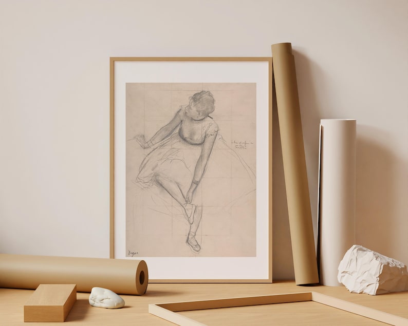 May include: Framed pencil sketch of a ballerina adjusting her shoe, with a light beige background. The artwork is in a gold frame. Rolls of paper and a wooden frame are on a light wood surface.