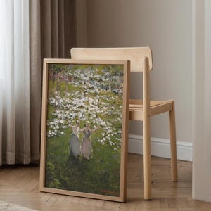 Op de afbeelding: Een ingelijst schilderij van drie vrouwen die in een veld met witte bloemen staan. Het schilderij is in een licht houten lijst en hangt aan een muur.