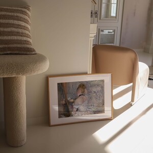 May include: A framed painting of a woman in a white dress, sitting in a chair. The painting is set against a light background with a soft, muted color palette. The painting is framed in a simple, wooden frame.
