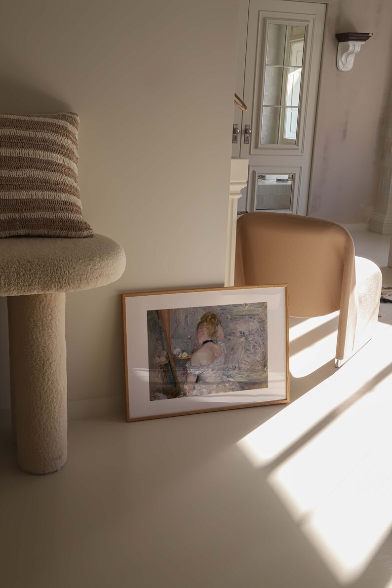 May include: A framed painting of a woman in a white dress, sitting in a chair. The painting is set against a light background with a soft, muted color palette. The painting is framed in a simple, wooden frame.