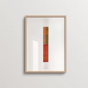 May include: A framed vertical textile art piece featuring a narrow, rectangular woven panel with a split color design. The left side displays orange and pink geometric patterns, while the right side shows green and brown tones, with loose threads.