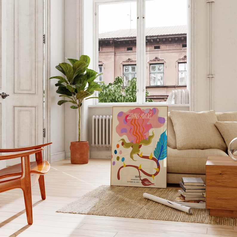May include: A colorful abstract art print with the text "Zhang Huanxi" and "Exhibition Room" in a white frame. The print is hanging on a white wall in a living room with a beige couch, a wooden coffee table, and a potted plant.