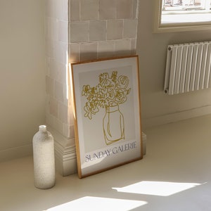 May include: A framed print with a minimalist line drawing of a bouquet of flowers in a vase. The drawing is in gold and the text "SUNDAY GALERIE" is in blue at the bottom of the print.