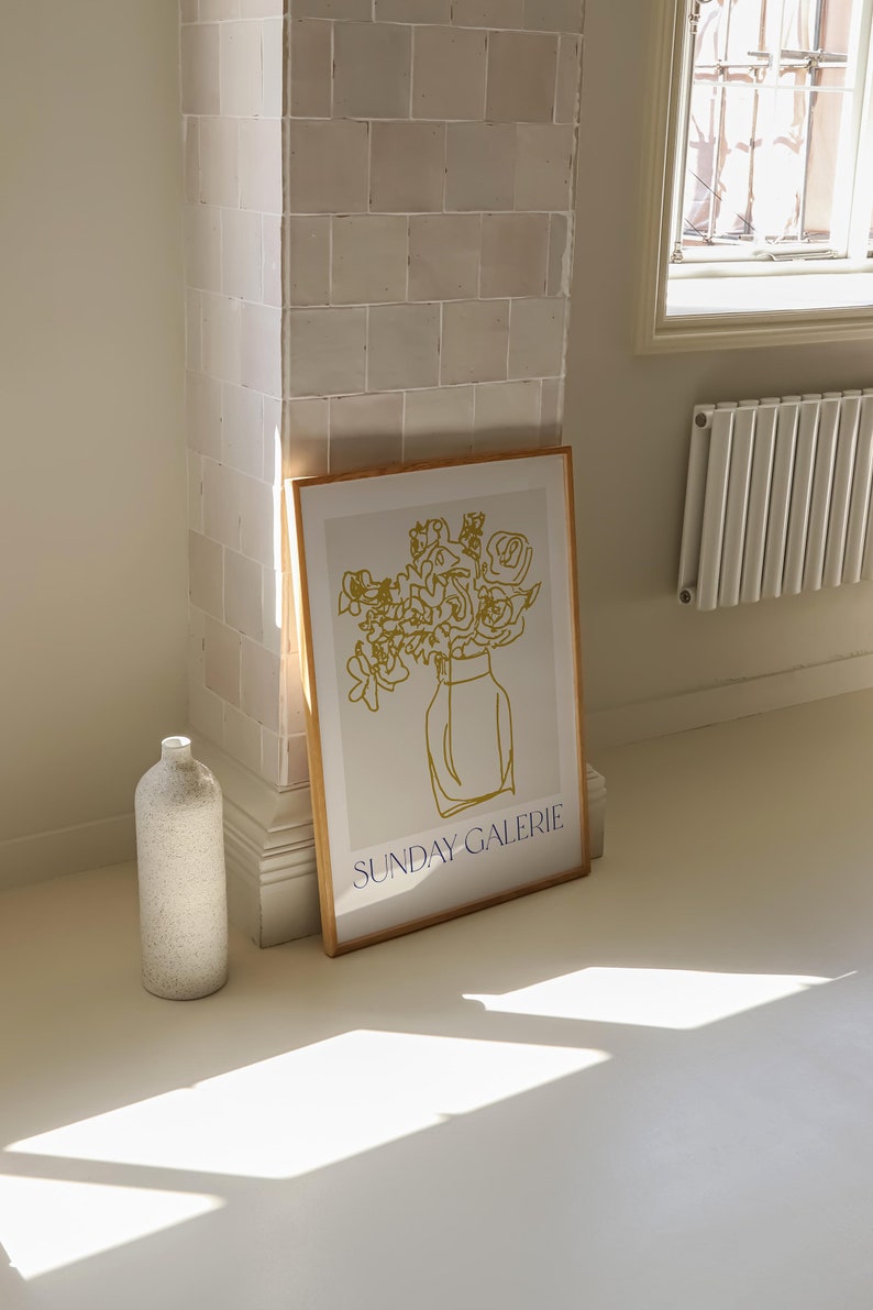 May include: A framed print with a minimalist line drawing of a bouquet of flowers in a vase. The drawing is in gold and the text "SUNDAY GALERIE" is in blue at the bottom of the print.