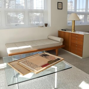 May include: A white tufted daybed with a wooden frame and a beige throw pillow sits in a modern living room with a glass coffee table and a wooden cabinet with drawers. A framed poster with the text "KINTAIL THANIG" is on the coffee table.