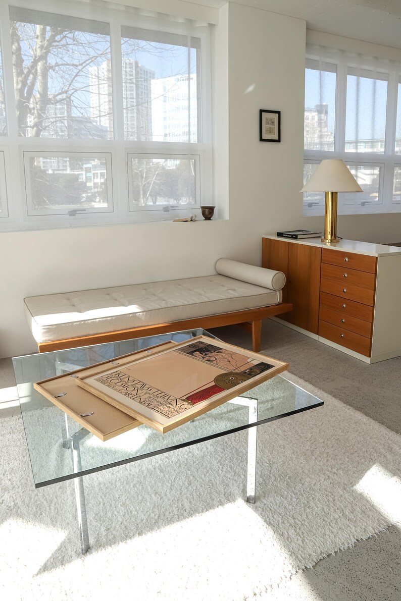 May include: A white tufted daybed with a wooden frame and a beige throw pillow sits in a modern living room with a glass coffee table and a wooden cabinet with drawers. A framed poster with the text "KINTAIL THANIG" is on the coffee table.