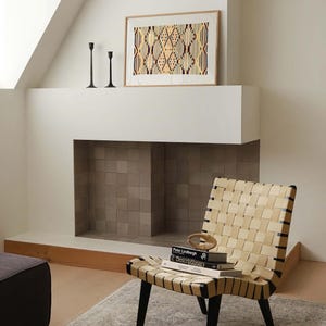K&ouml;nnte beinhalten: Ein Stuhl aus geflochtenem Leder mit schwarzen Beinen steht vor einem Kamin. Ein gerahmtes Kunstwerk mit einem geometrischen Muster h&auml;ngt &uuml;ber dem Kamin. Zwei schwarze Kerzenhalter befinden sich auf dem Kaminsims. B&uuml;cher und ein dekoratives Objekt liegen auf dem Stuhl.