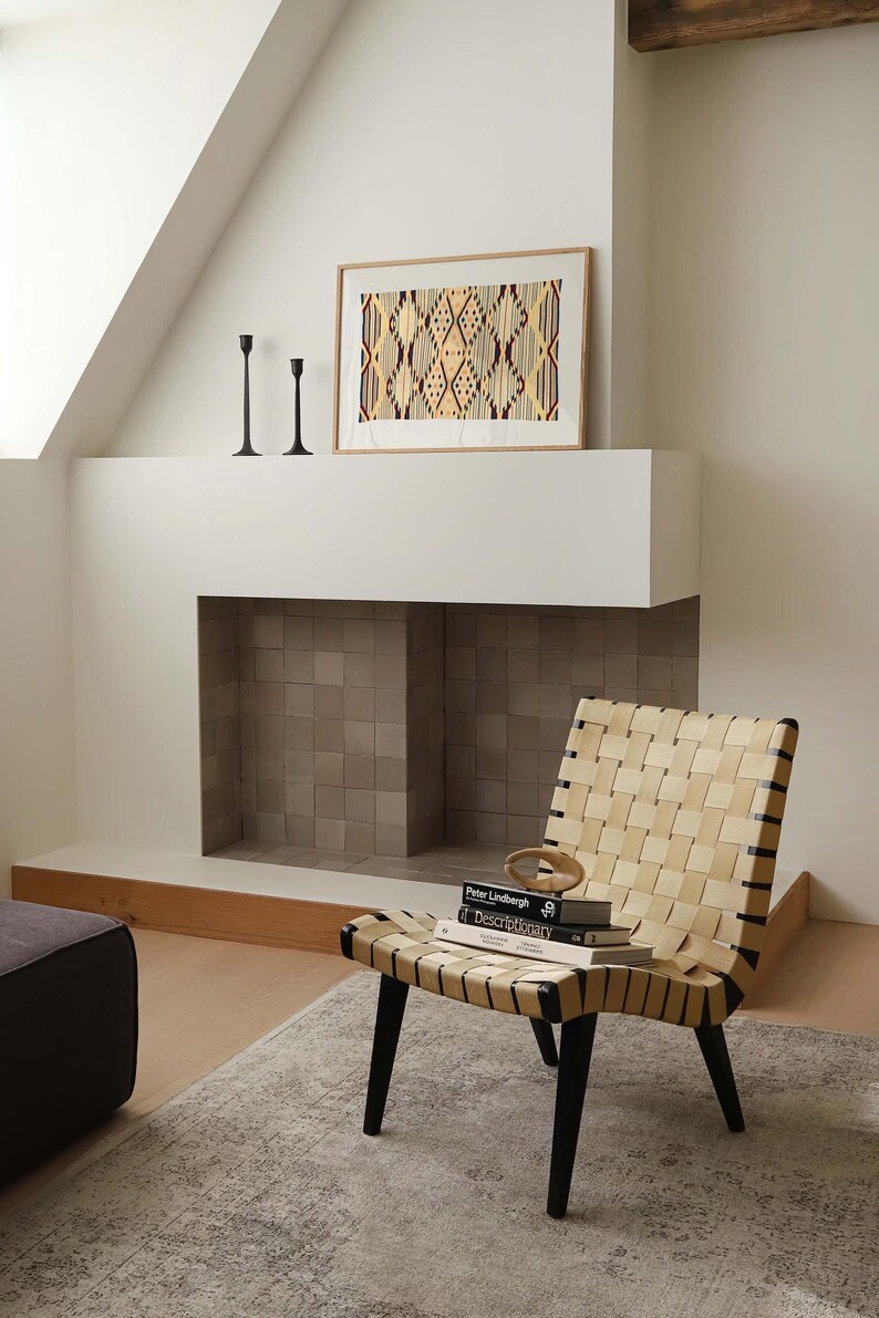 K&ouml;nnte beinhalten: Ein Stuhl aus geflochtenem Leder mit schwarzen Beinen steht vor einem Kamin. Ein gerahmtes Kunstwerk mit einem geometrischen Muster h&auml;ngt &uuml;ber dem Kamin. Zwei schwarze Kerzenhalter befinden sich auf dem Kaminsims. B&uuml;cher und ein dekoratives Objekt liegen auf dem Stuhl.