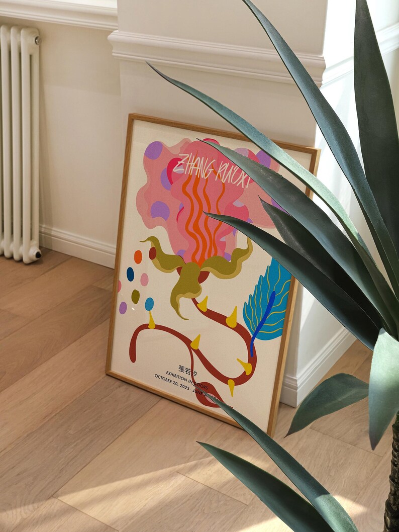 May include: A framed poster with a colorful abstract design featuring a pink, orange, and yellow flower with blue and green accents. The poster has the text "ZHANG KUOXI" and "EXHIBITION IN PROGRESS" with the dates "OCTOBER 20, 2023 - JUNE 2024".
