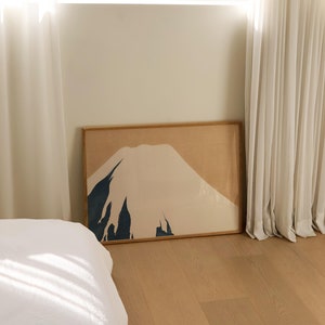 以下が含まれることがあります： ベージュ、ブラウン、ブルーのミニマルな山岳風景の額装されたプリント。プリントは、白いカーテンと白いベッドのある寝室の白い壁にかけられています。
