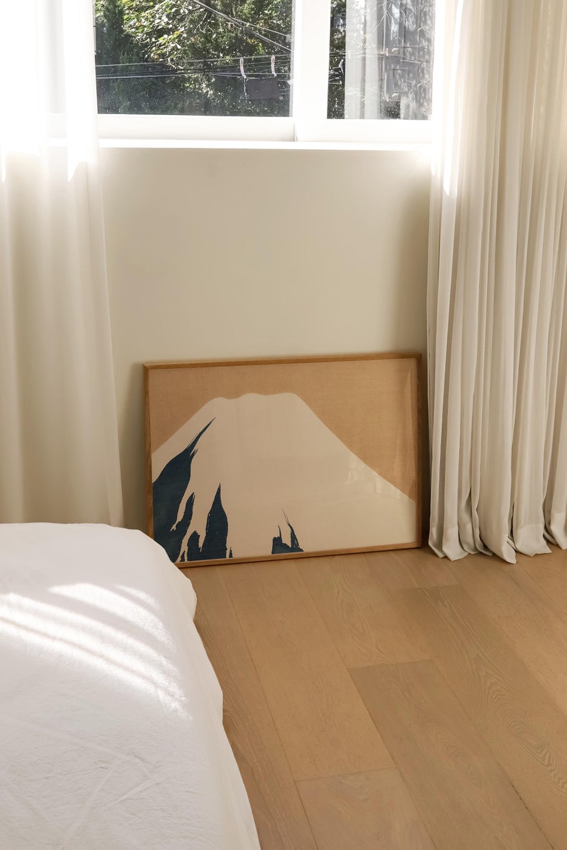 以下が含まれることがあります： ベージュ、ブラウン、ブルーのミニマルな山岳風景の額装されたプリント。プリントは、白いカーテンと白いベッドのある寝室の白い壁にかけられています。