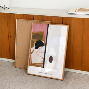 May include: A framed print of a painting by Onchi Koshiro, a Japanese artist. The painting depicts a woman in a pink and gold room. The print is in a wooden frame and is hanging on a wall.
