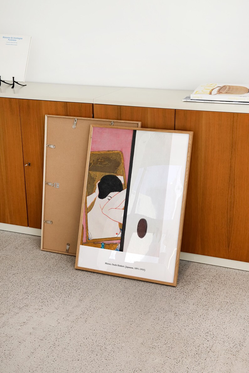 May include: A framed print of a painting by Onchi Koshiro, a Japanese artist. The painting depicts a woman in a pink and gold room. The print is in a wooden frame and is hanging on a wall.