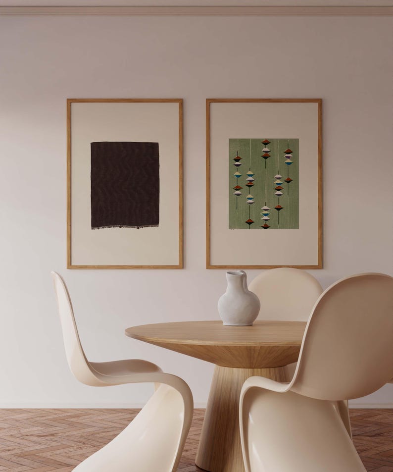 May include: Two framed abstract art prints with wooden frames. One print features a dark brown rectangular shape, and the other has a green background with a pattern of hanging shapes. A round wooden table and three cream-colored chairs are in the foreground.