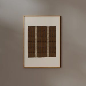 Puede incluir: Obra de arte enmarcada que presenta un textil marr&oacute;n con un patr&oacute;n de cuadr&iacute;cula y dos rayas blancas verticales. La obra de arte est&aacute; enmarcada en un marco dorado y montada sobre un fondo blanco. La pieza tiene una est&eacute;tica r&uacute;stica y artesanal.