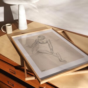 May include: A framed pencil sketch of a seated figure, displayed on a wooden surface. A white ceramic lamp and a mug are also on the surface. The artwork is in a light wood frame. The scene is lit by natural light.