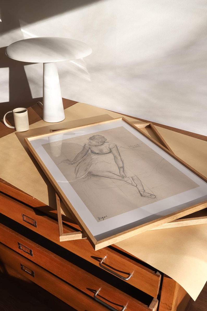 May include: A framed pencil sketch of a seated figure, displayed on a wooden surface. A white ceramic lamp and a mug are also on the surface. The artwork is in a light wood frame. The scene is lit by natural light.