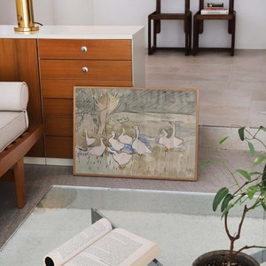 May include: A rectangular framed art print featuring a watercolor illustration of a group of white geese in a grassy field with a tree. The artwork is enclosed in a light-colored wooden frame and displays a signature in the bottom right corner.