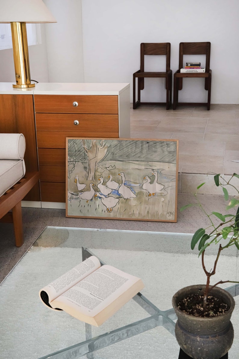May include: A rectangular framed art print featuring a watercolor illustration of a group of white geese in a grassy field with a tree. The artwork is enclosed in a light-colored wooden frame and displays a signature in the bottom right corner.