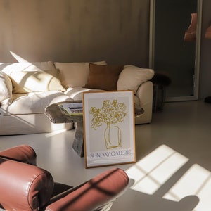 May include: A white couch with brown pillows in a room with a brown leather chair and a framed print of a line drawing of flowers in a vase. The print has the text "SUNDAY GALERIE" at the bottom.