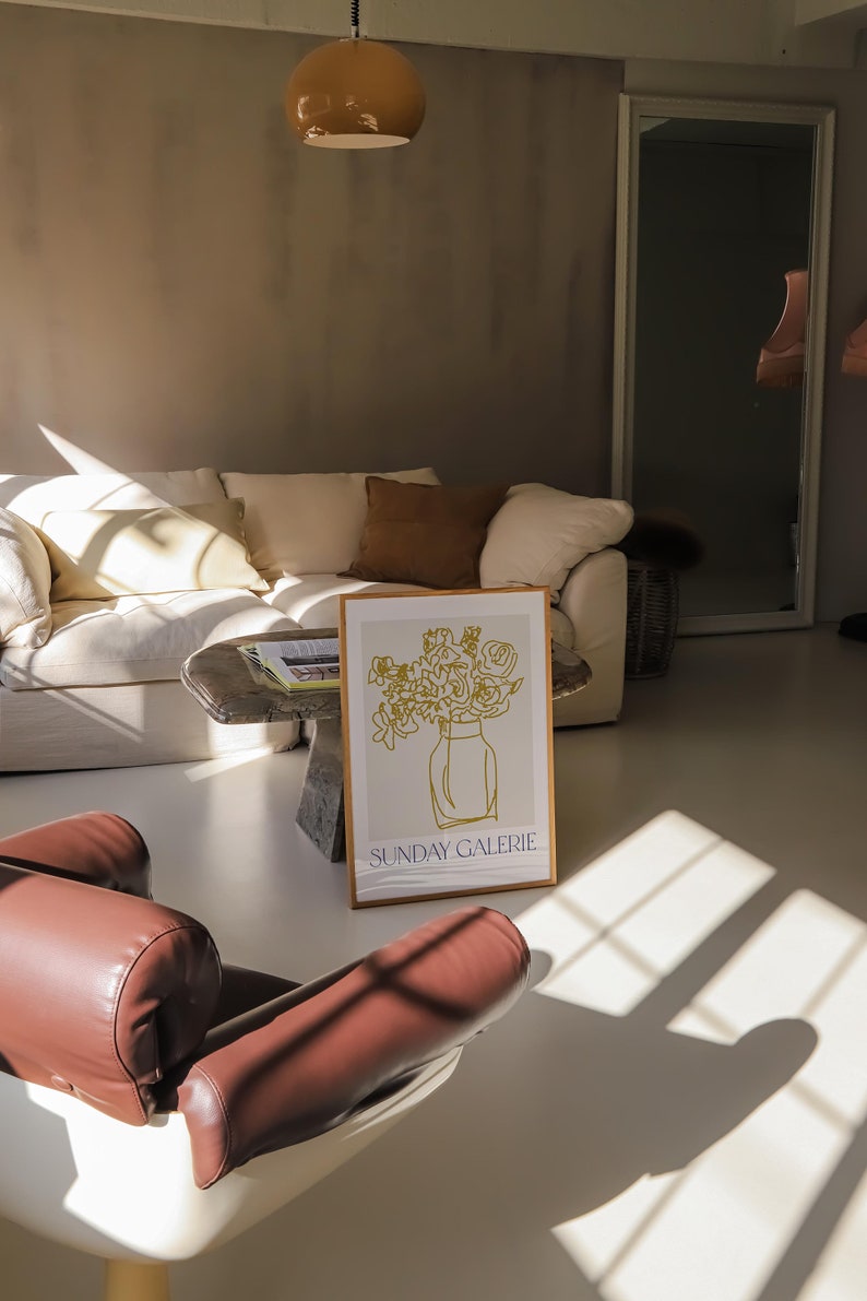 May include: A white couch with brown pillows in a room with a brown leather chair and a framed print of a line drawing of flowers in a vase. The print has the text "SUNDAY GALERIE" at the bottom.