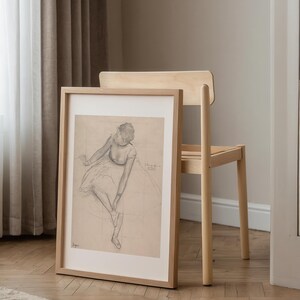 May include: A framed pencil sketch of a seated figure in a light wood frame. The artwork is leaning against a light wood chair. The sketch is on a grid background, and the figure is wearing a dress.