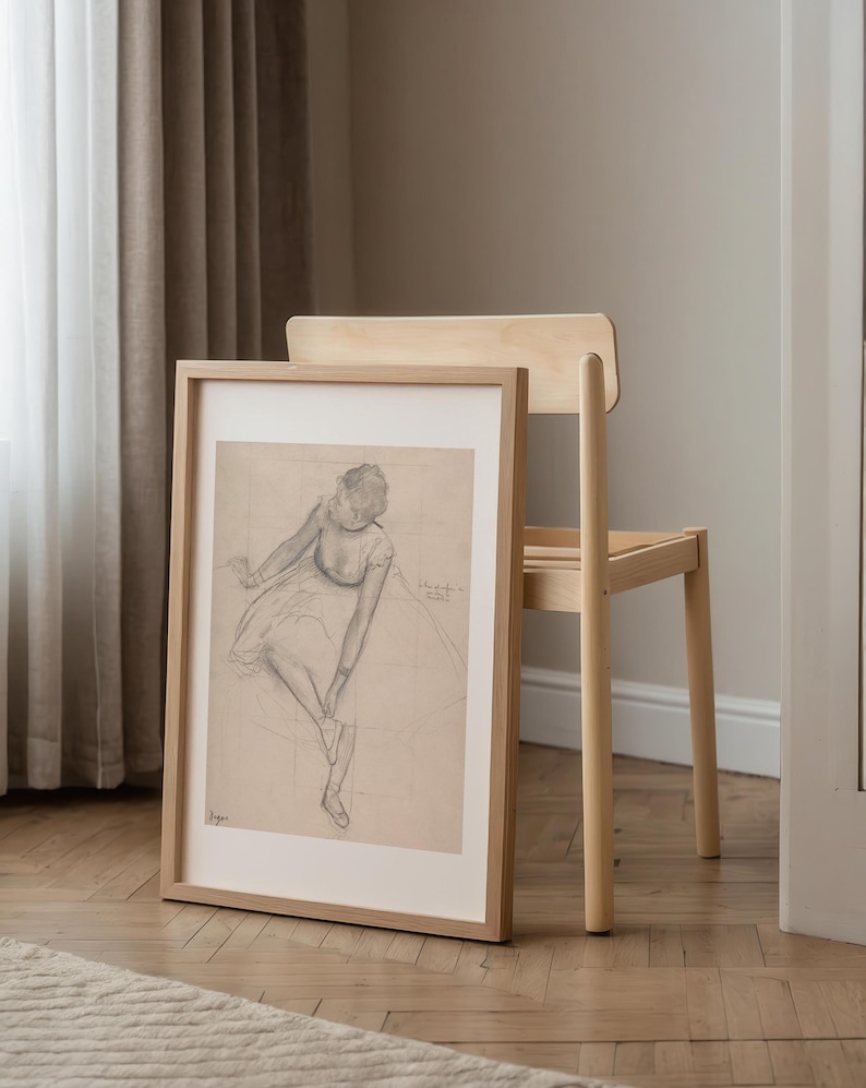 May include: A framed pencil sketch of a seated figure in a light wood frame. The artwork is leaning against a light wood chair. The sketch is on a grid background, and the figure is wearing a dress.