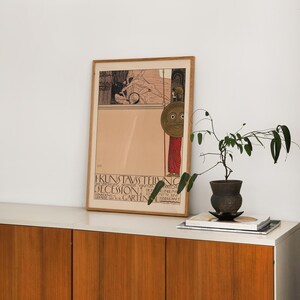 May include: A framed vintage poster with a black and white illustration of a woman holding a shield with a face on it. The poster is titled "I. Kunstausstellung der Vereinigung bildender K&uuml;nstler &Ouml;sterreichs Secession" and is for an exhibition in the "Gartenbau Gesellschaft Wien" in 1900.